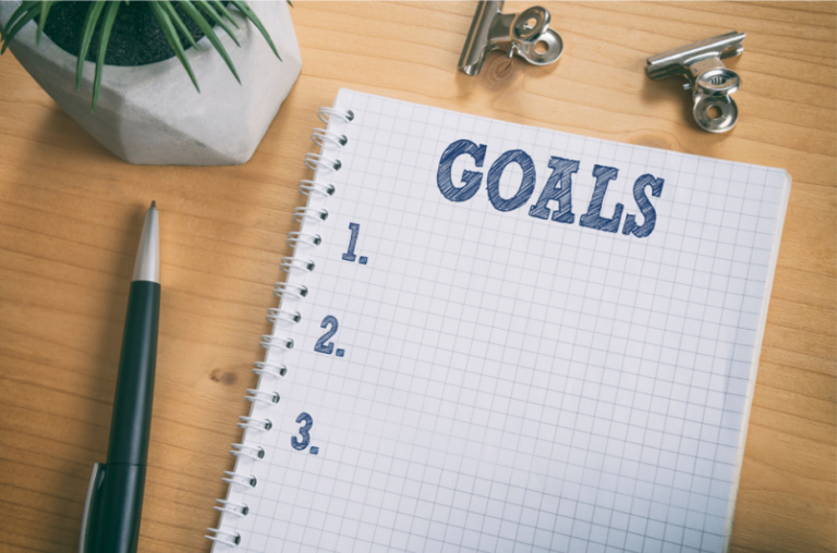 A spiral notebook with a grid pattern lies open on a wooden surface, displaying a "Goals" title with three numbered bullet points beneath it. Nearby are a pen, a plant, and metal clips.